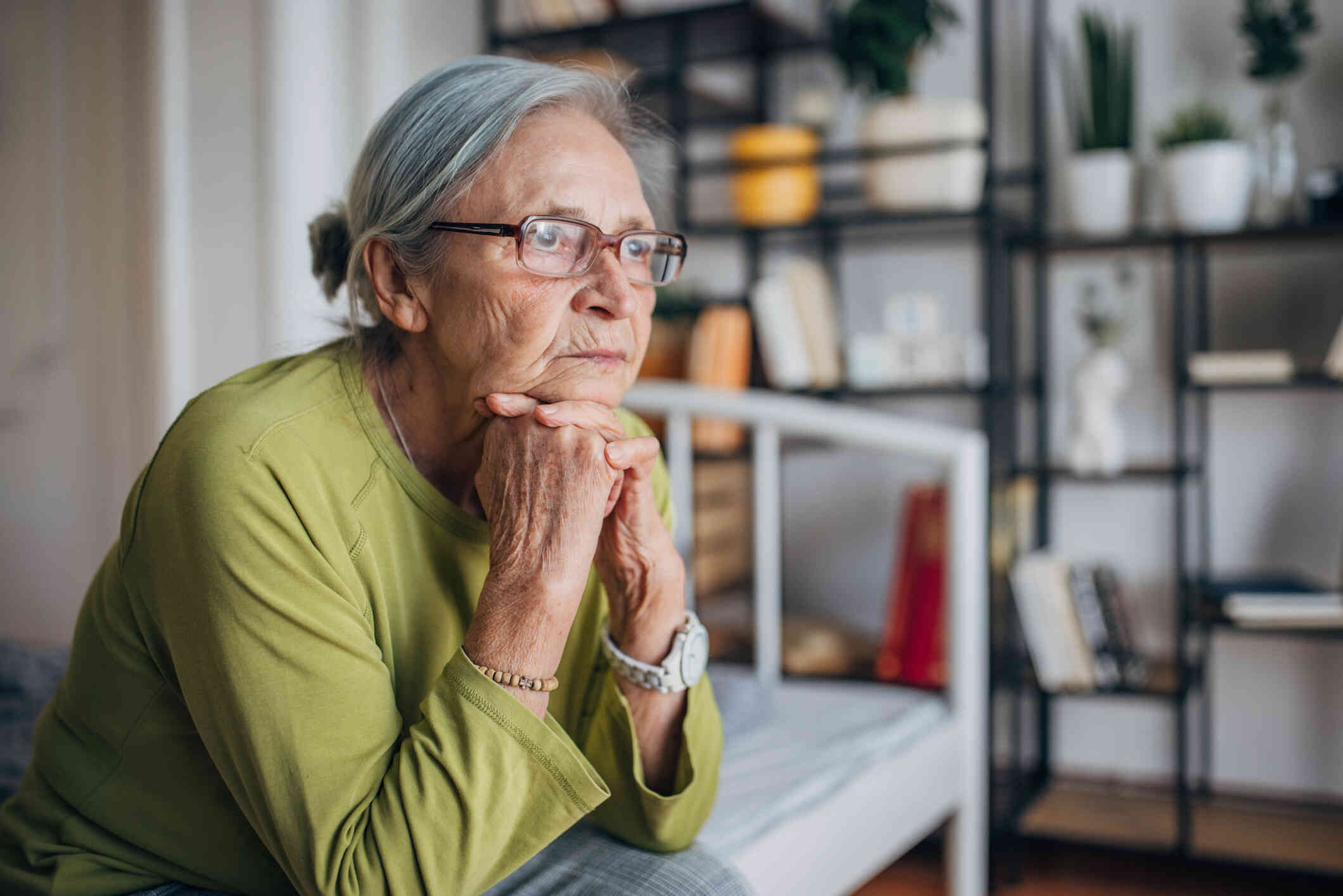 A close up of an elderly woman in a green shirt sits hunched over on the bed and sadly rests her head on her hands while gazing off.